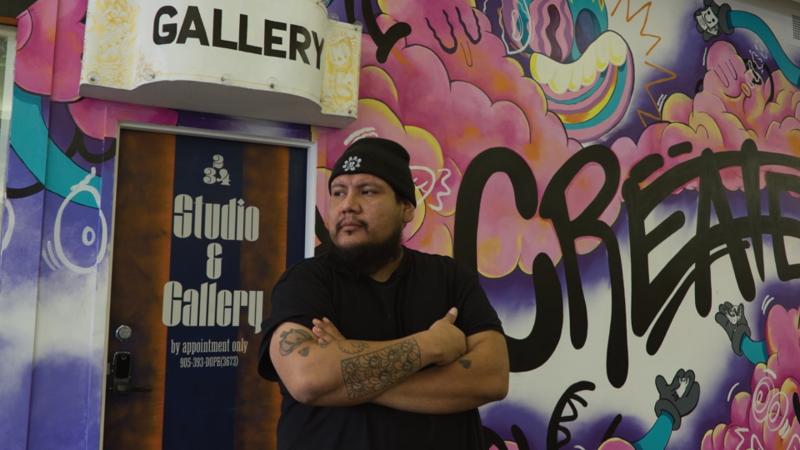 Dope Chief standing infront of his studio space, with a colorful mural behind him.