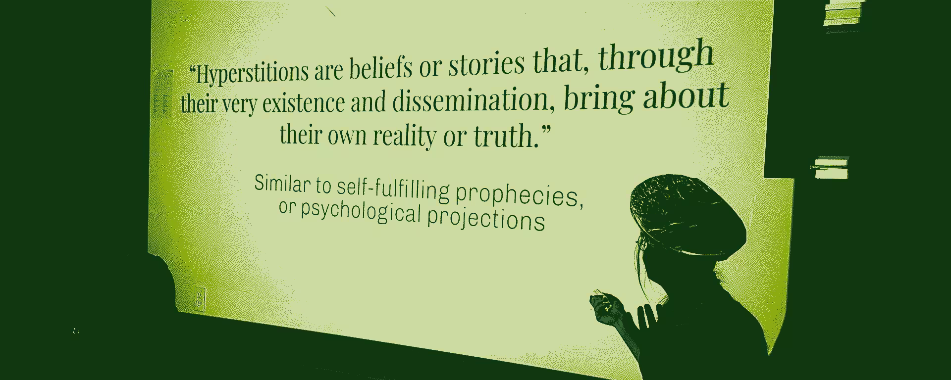 Teacher infront of projection screen that displays this quote: Hyperstitions are beliefs or stories that, through their very existence and dissemination, bring about their own reality or truth - similar to self-fullfilling prophecies, or psychological projections.