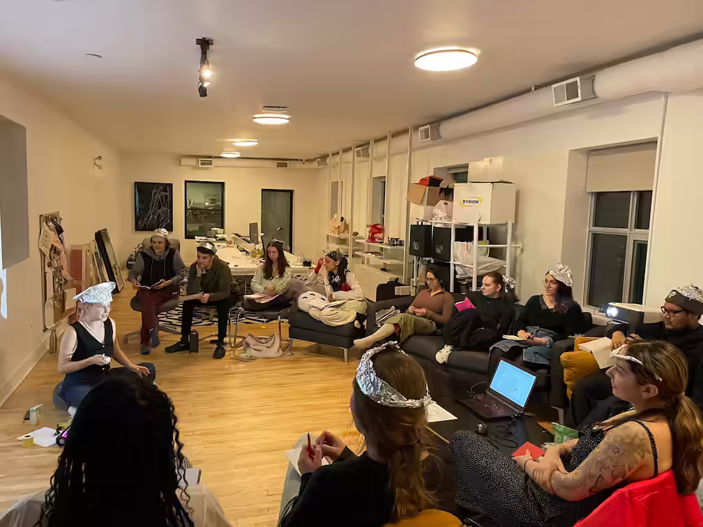 Photograph of poetry students sitting in a half-circle, wearing aluminum hats, listening to lecturer in the middle.